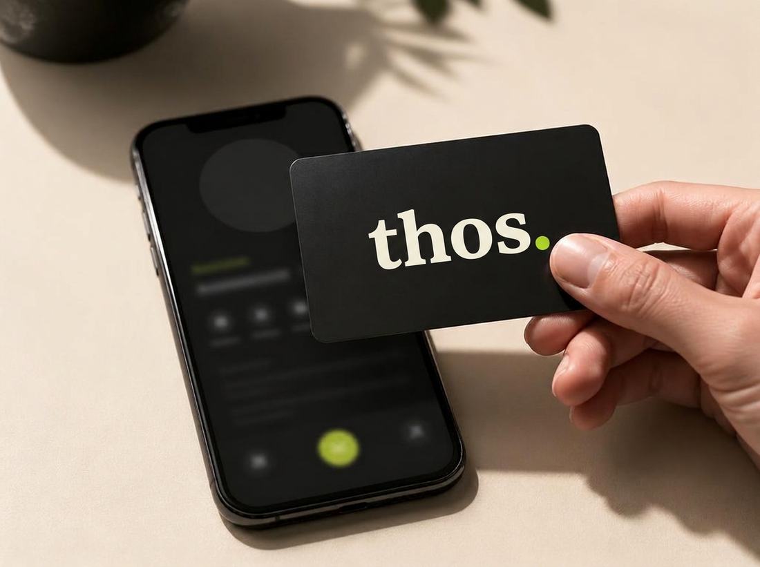 A hand tapping a matte black NFC business card against a phone, on a warm bone surface.